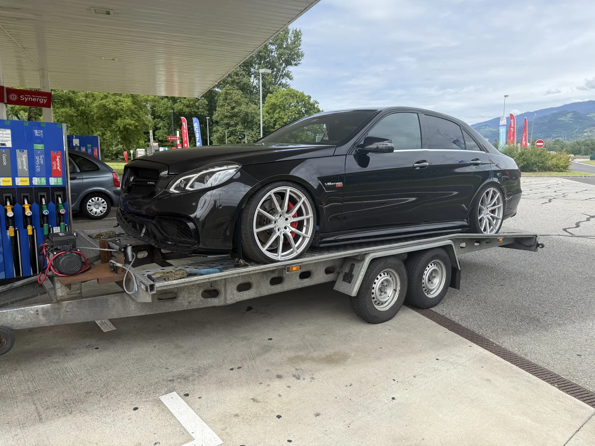 Berline sportive noire aux jantes argentées chargée sur une remorque porte-voiture à double essieu, dans une station-service
