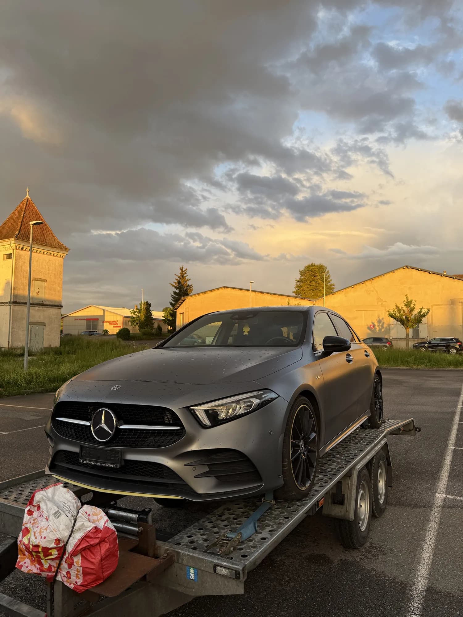 Compacte Mercedes gris mat arrimée sur une remorque porte-voiture, sur un parking mouillé au coucher du soleil