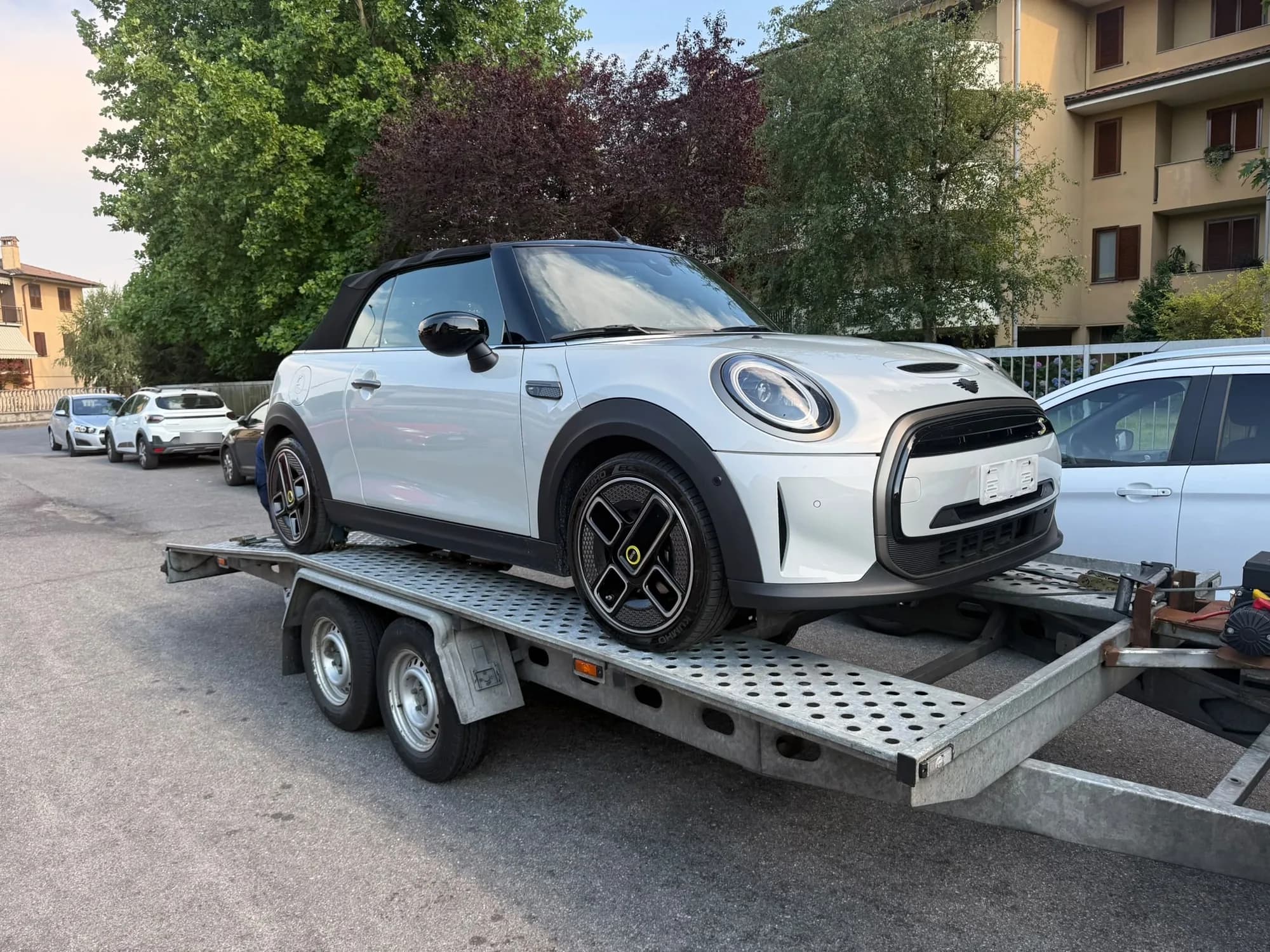 Petit cabriolet blanc à capote noire chargé sur une remorque porte-voiture galvanisée, dans une rue résidentielle bordée d'arbres