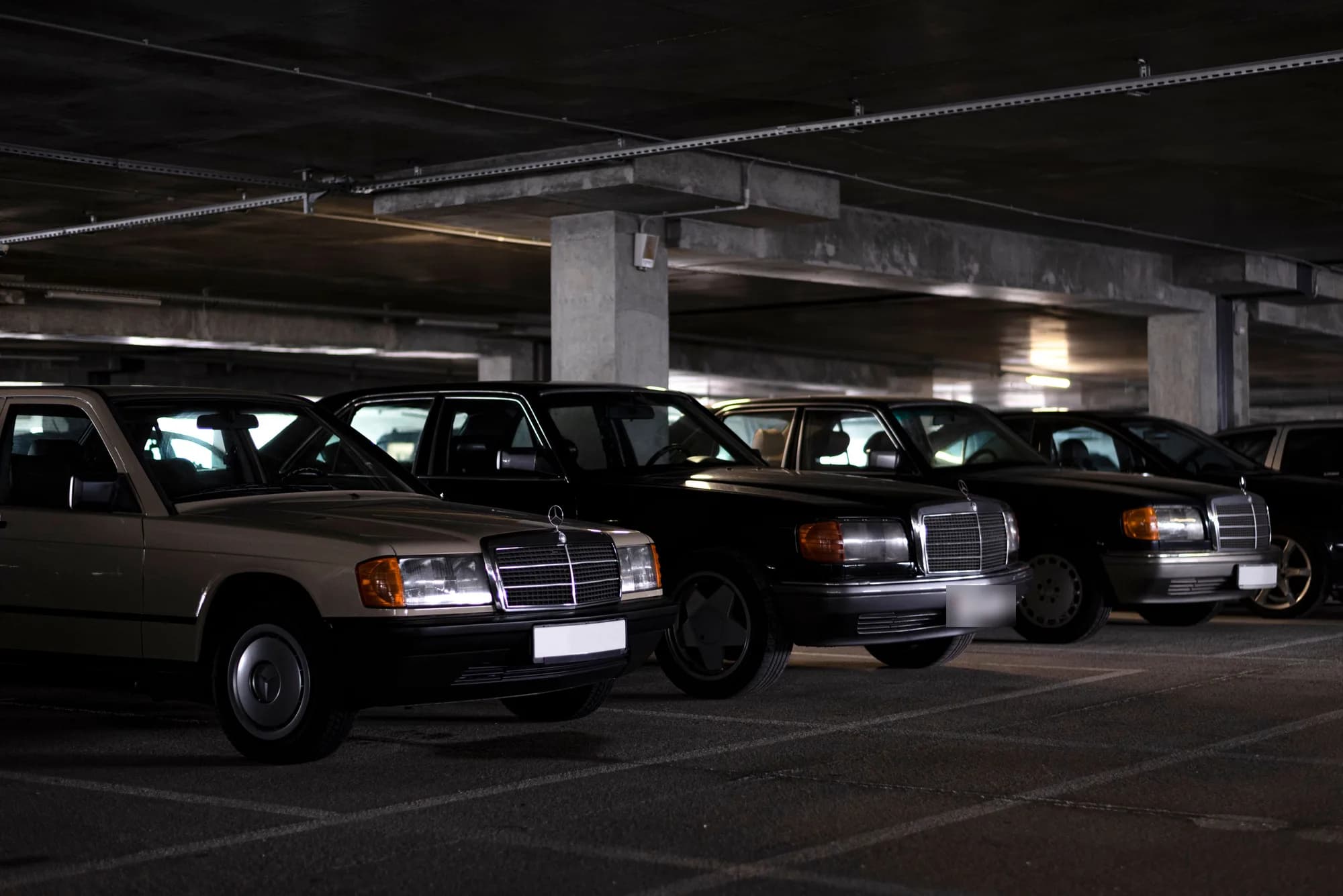 Berlines Mercedes anciennes, une beige et les autres noires, garées côte à côte dans un parking couvert en béton faiblement éclairé