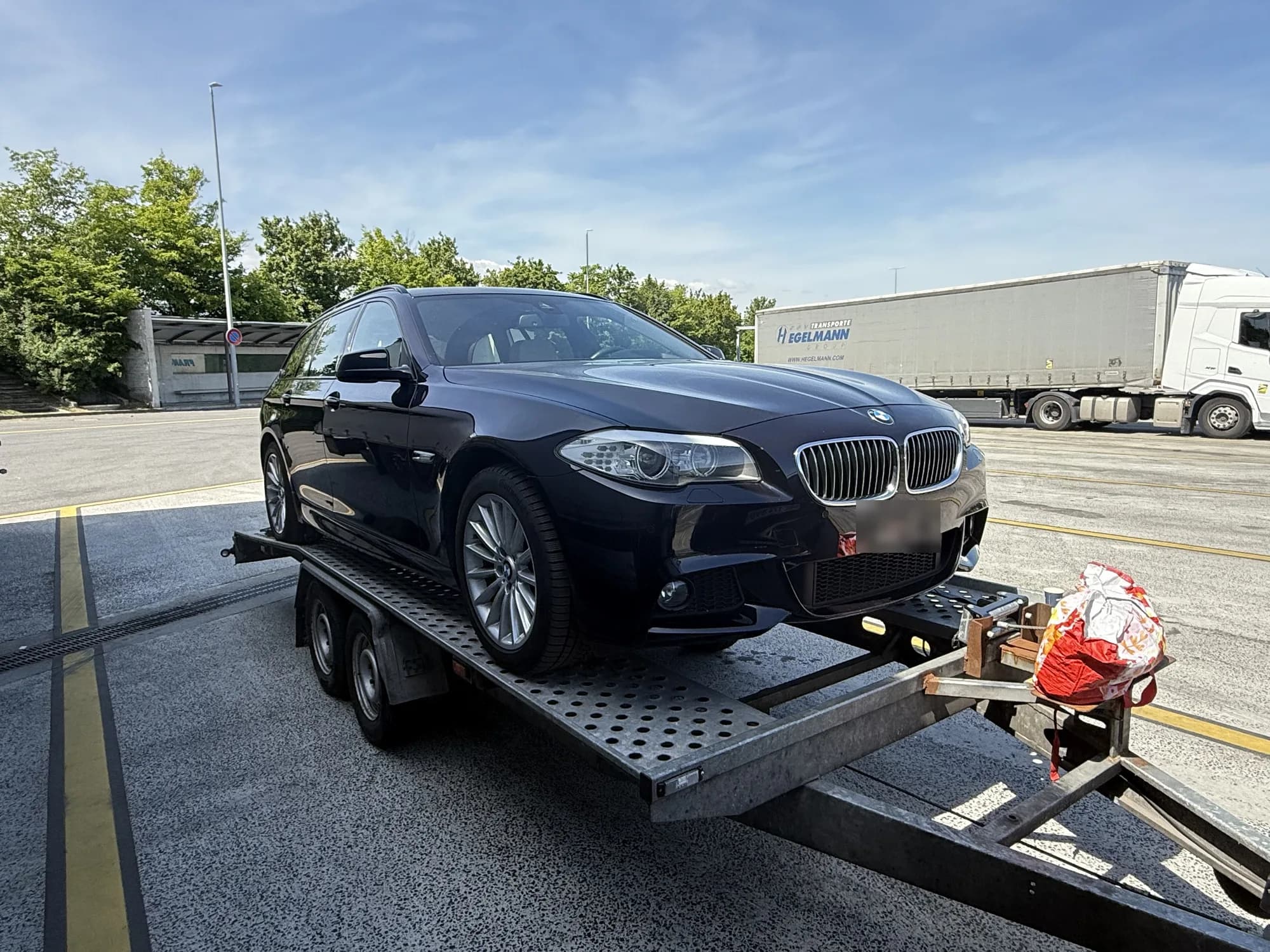 Break BMW noir chargé sur une remorque porte-voiture à plateau perforé, sur un vaste parking avec un semi-remorque en arrière-plan